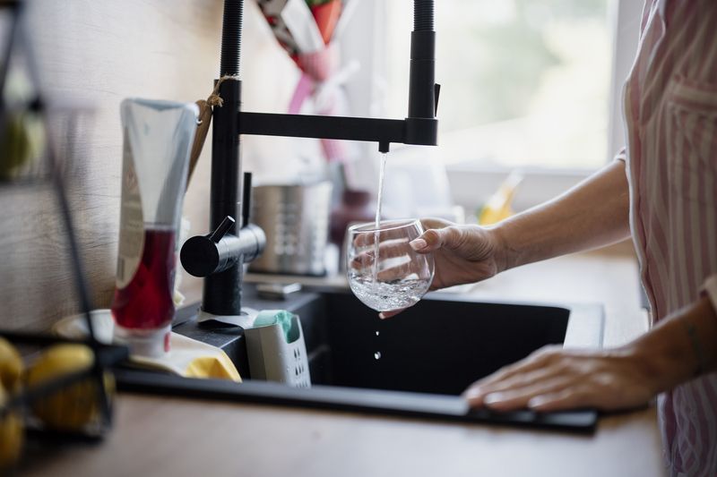 Hand filling a clear drinking glass from a black kitchen faucet over the sink